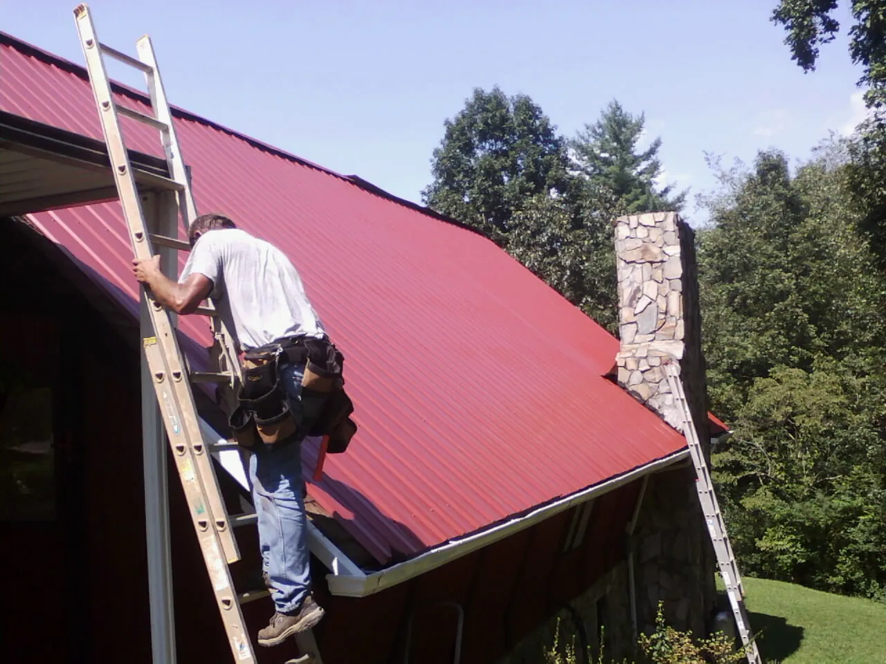 Roofer on a ladder beside a red metal roof and stone chimney — Soffit & Fascia Repair by Roofing Brothers in Bellefontaine